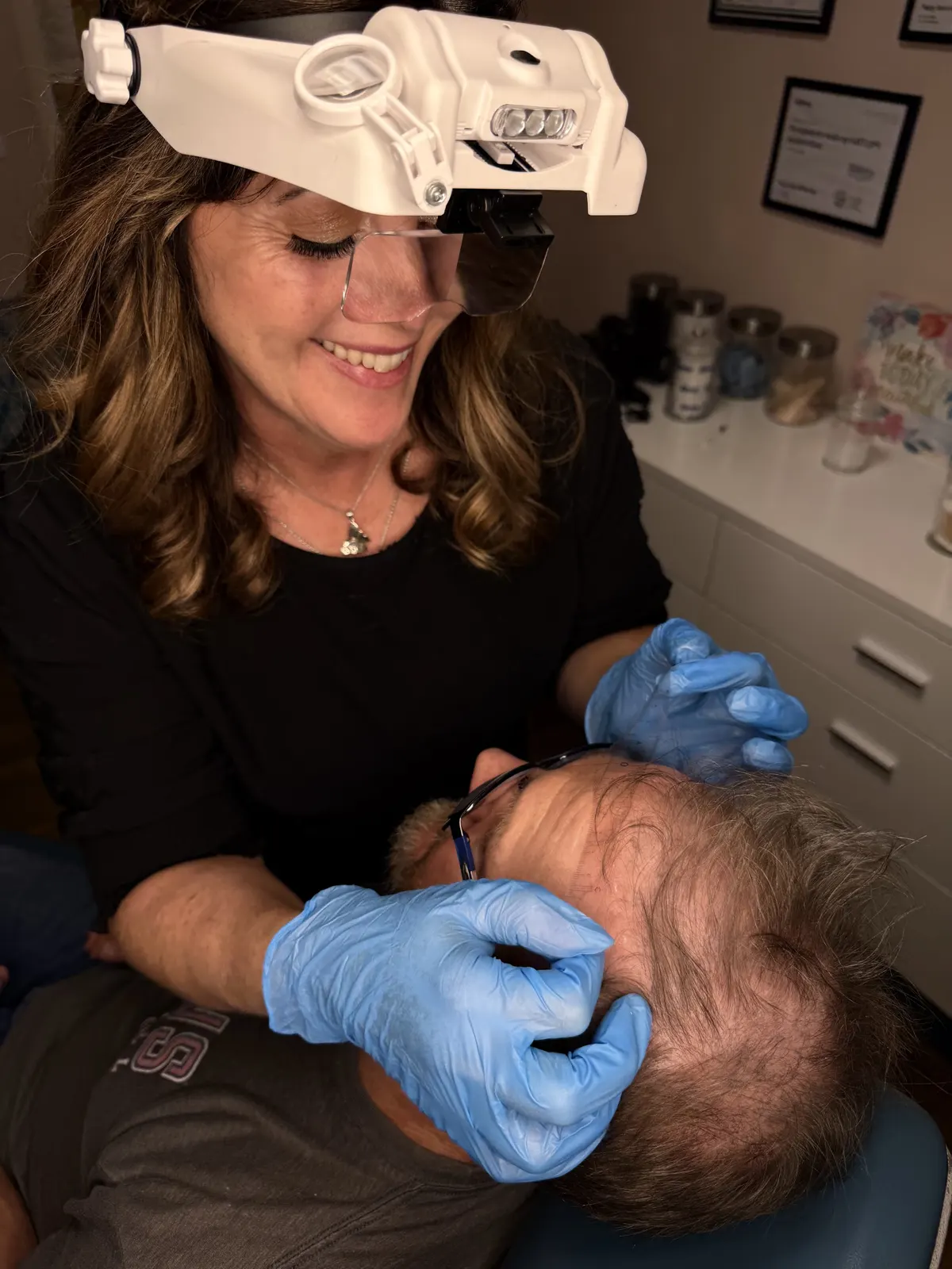 Tracy Whitman smiling as she works on a client's scalp under a magnifying headlamp